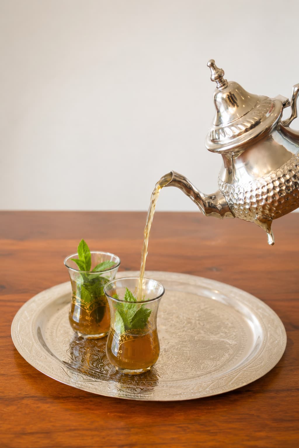 Moroccan mint tea being poured from height from an ornate silver teapot into two small traditional glasses with fresh mint sprigs, on an engraved silver tray on a wooden table