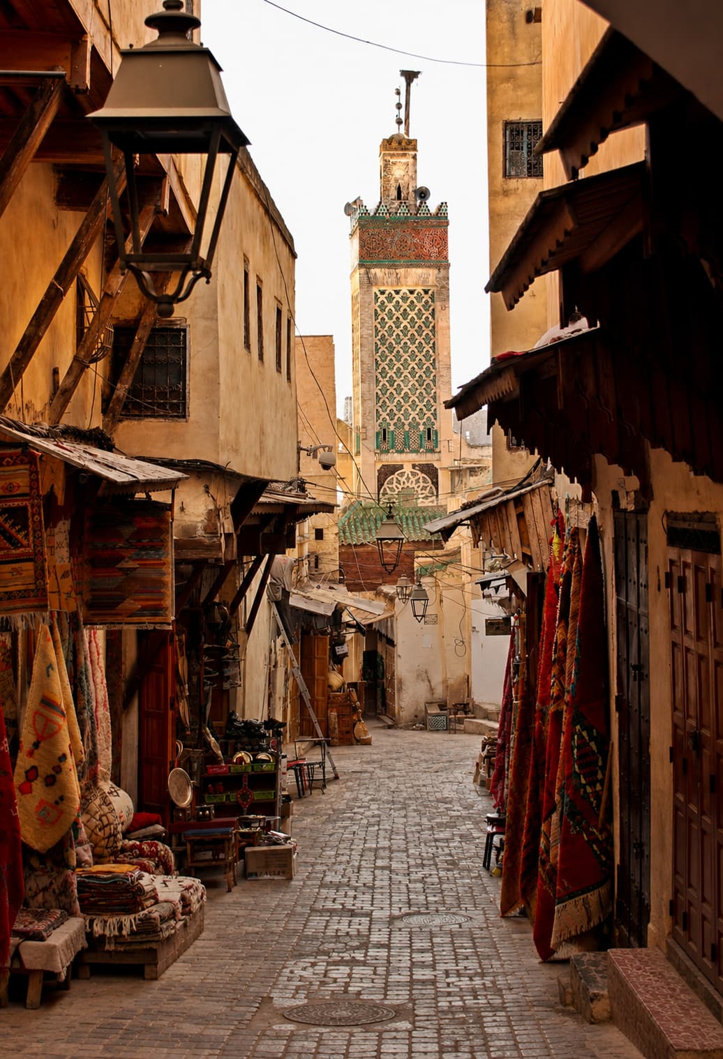 A souk alley in the Fes medina — carpet shops and lanterns lining a cobblestone street, an ornate green-tiled mosque minaret rising at the far end