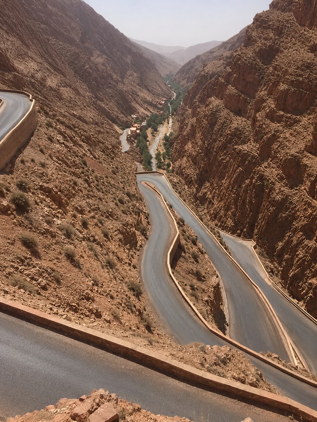 The dramatic switchback road descending through the Dadès Gorge in Morocco, with red-rock canyon walls on both sides and a ribbon of green valley below — part of the driving route toward Merzouga