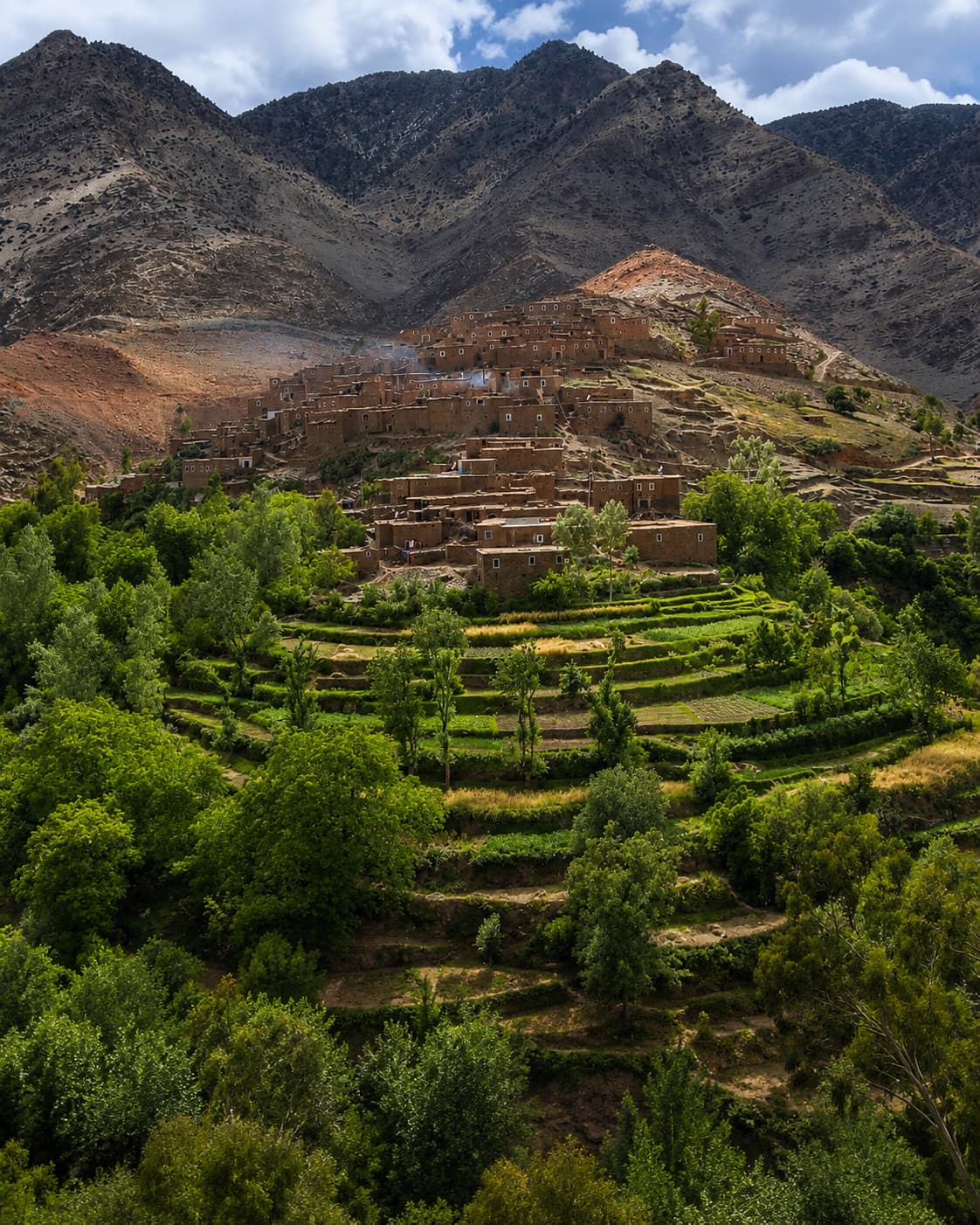 A traditional Berber village built into a steep hillside in the High Atlas Mountains of Morocco, surrounded by terraced green fields — the landscape between Marrakech and the desert on the route south