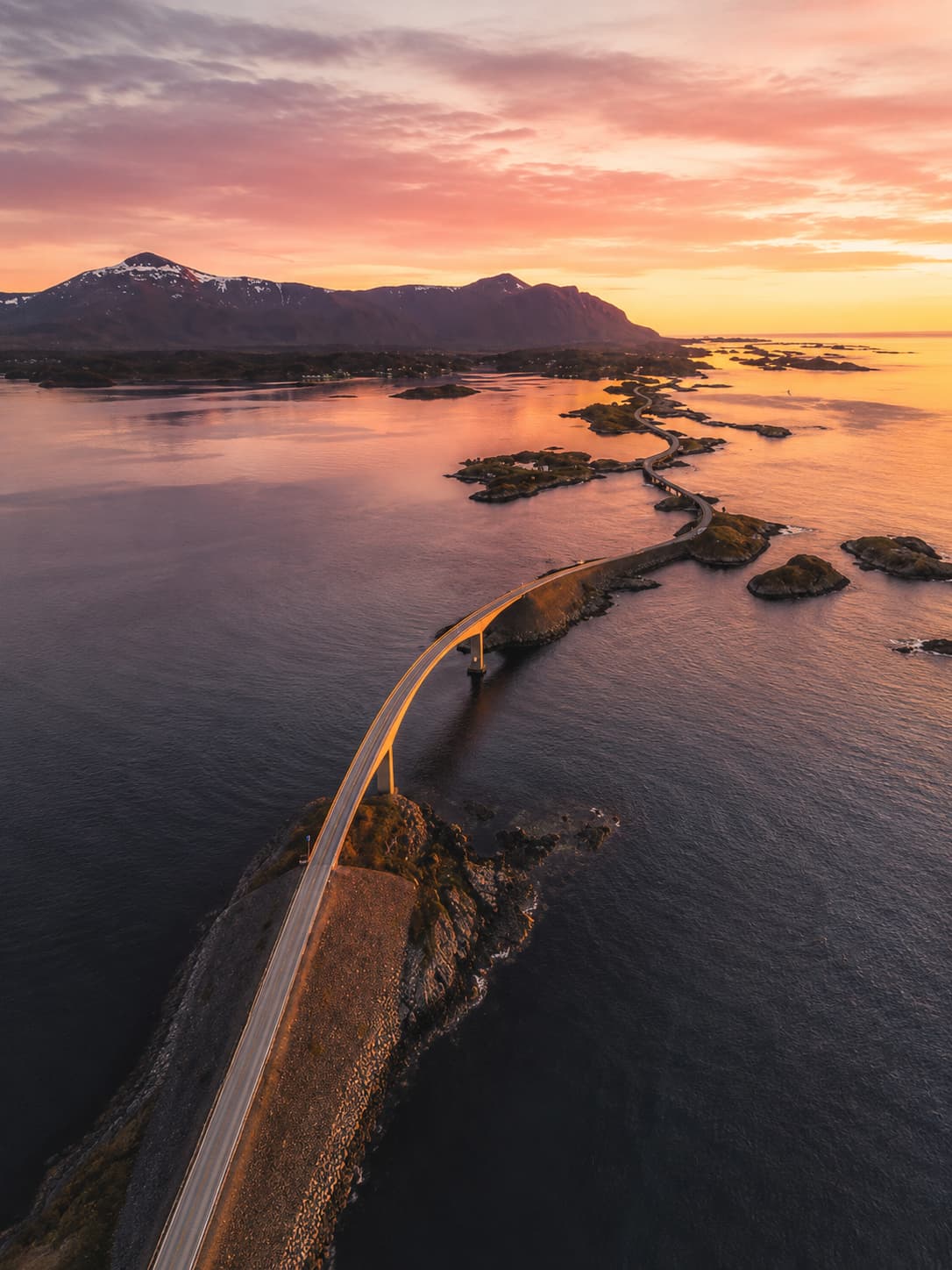 Aerial view of the Atlantic Ocean Road bridge chain at sunset, the Storseisundet Bridge glowing gold against a pink and orange sky, sea stretching to the horizon