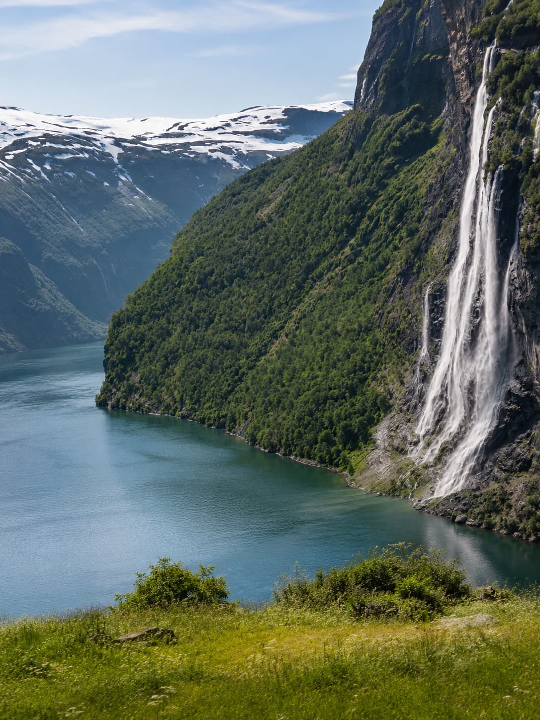 The Seven Sisters waterfall cascading down a steep green cliff into Geirangerfjord, snow-capped mountains in the background under a blue summer sky