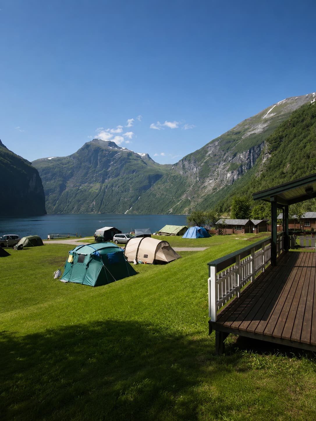 Campsite at the edge of Geirangerfjord, tents pitched on green grass with the fjord and dramatic mountain walls behind, a wooden cabin deck in the foreground