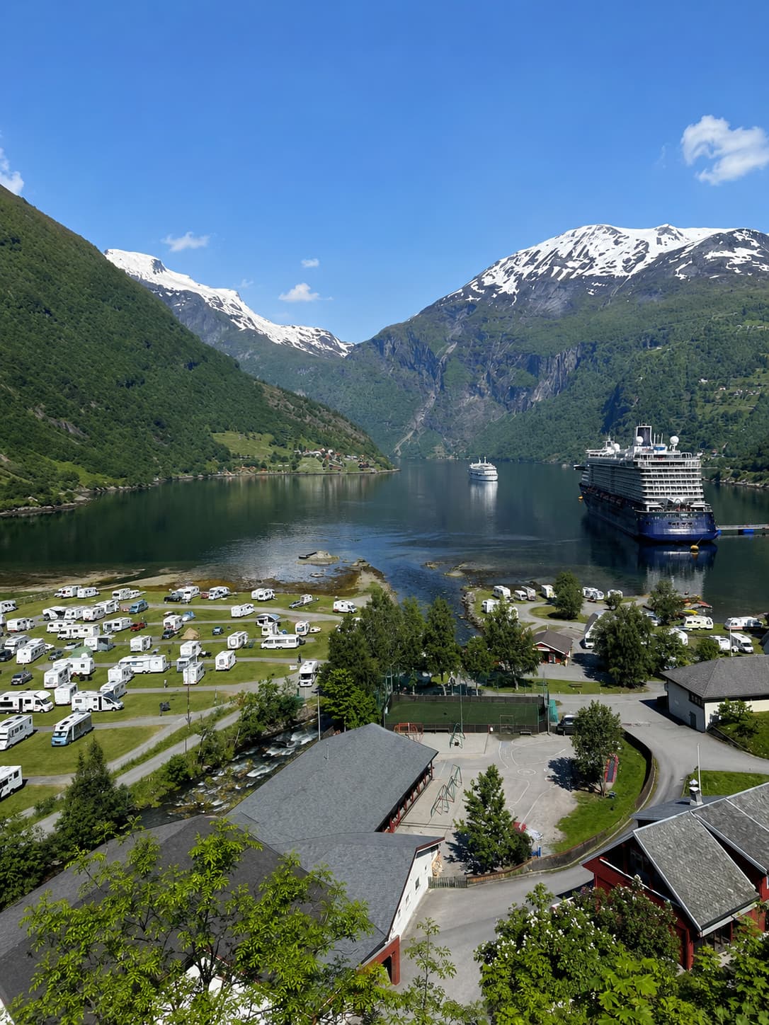 Geiranger village seen from above in summer, dozens of campervans parked on the waterfront and a large cruise ship anchored in the fjord, surrounded by steep green mountains with snow on the peaks