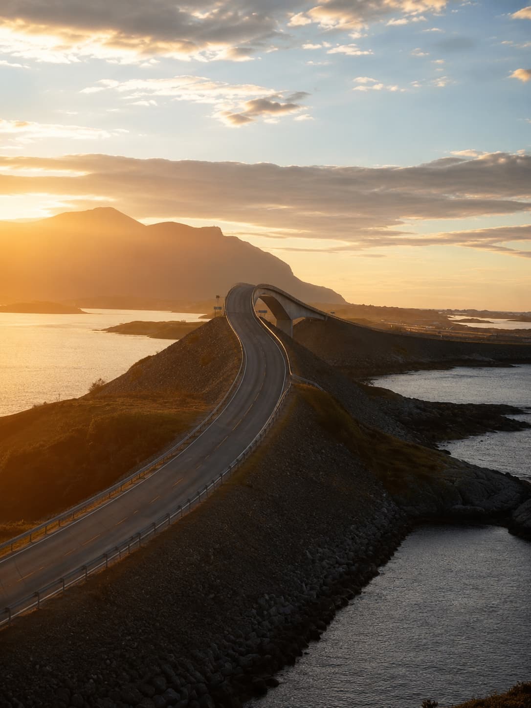 The Storseisundet Bridge on the Atlantic Ocean Road at golden sunset, the sweeping S-curve of the road lit in warm orange light, sea on both sides and a mountain silhouette behind