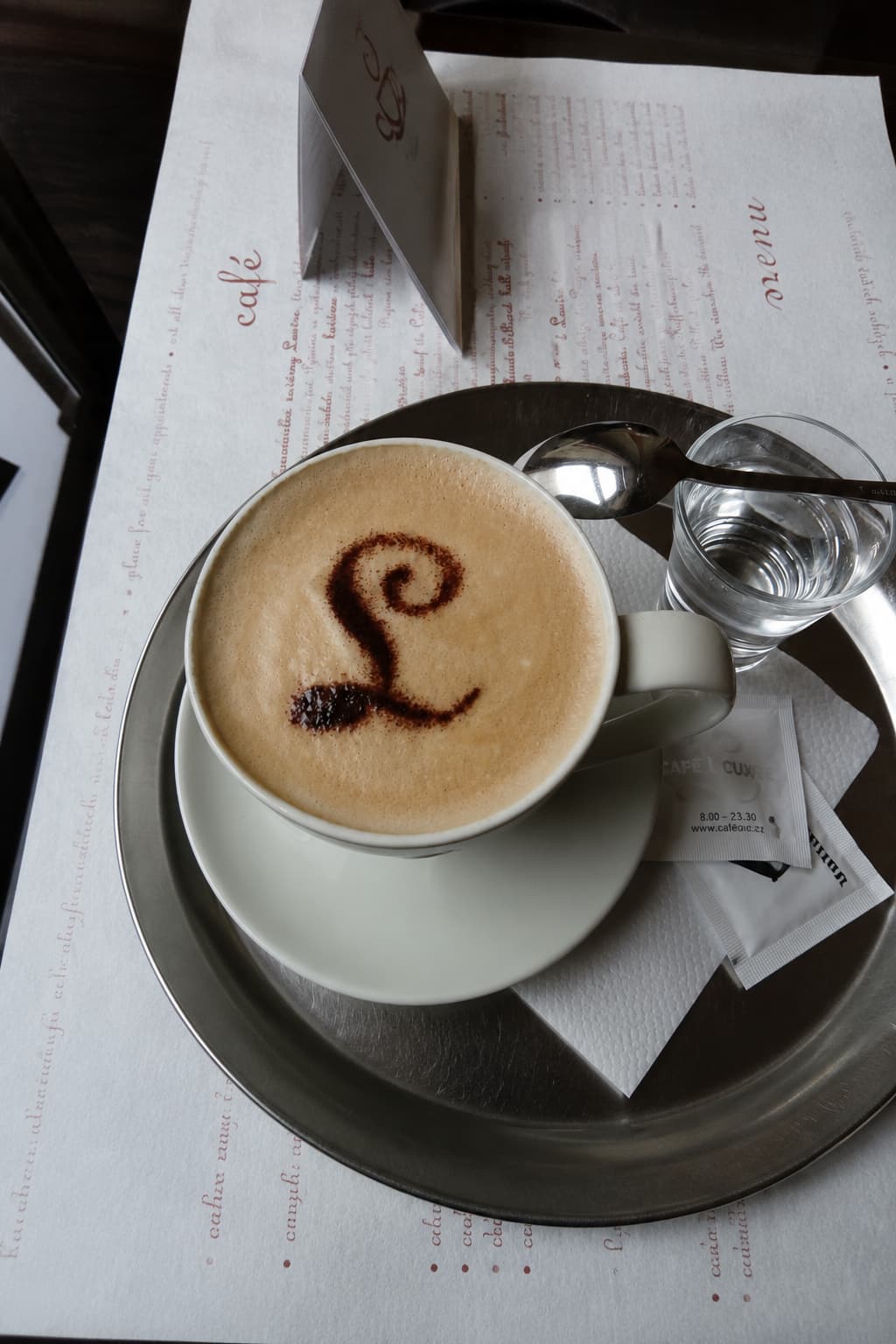 Cappuccino with the Café Louvre 'L' logo on the foam, served on a silver tray — one of the best cafes in Prague for atmosphere