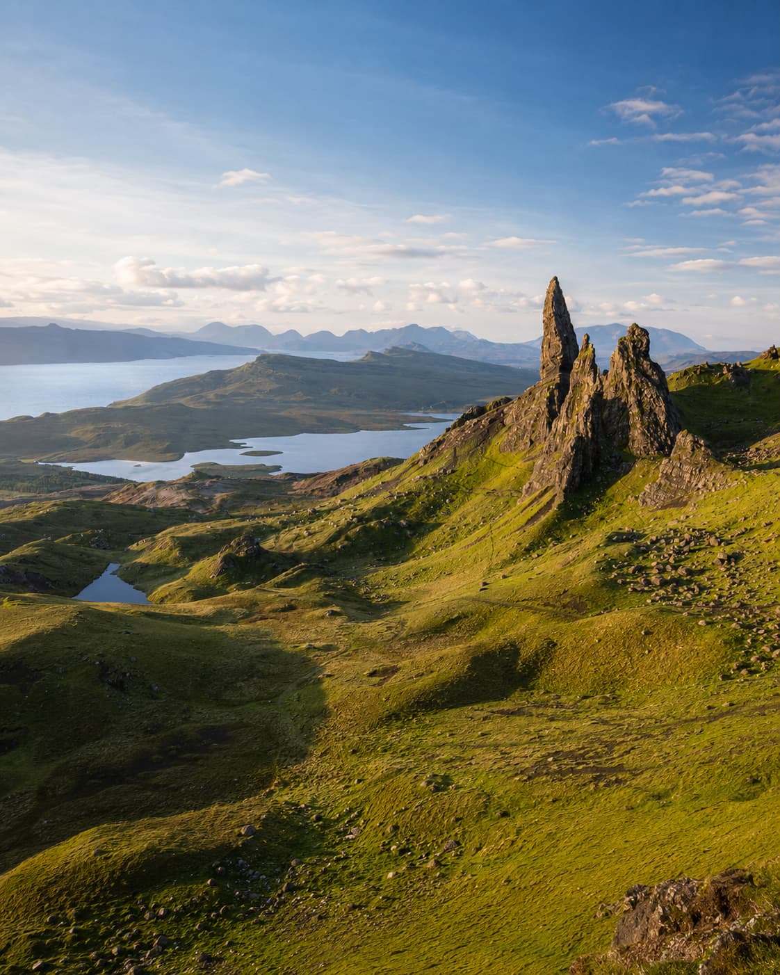 Old Man of Storr on the Isle of Skye — jagged basalt pinnacles rising above green moorland with sea lochs visible below, one of the highlights of any Scotland road trip itinerary