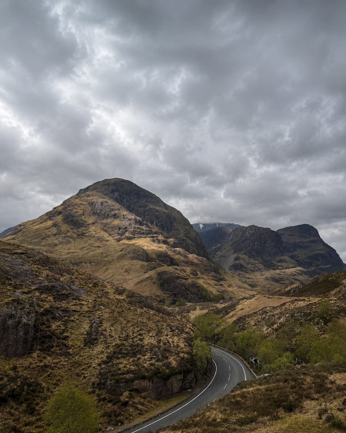 The road through Glencoe winding between steep mountains under a dramatic overcast sky — one of the most striking drives on any Scotland road trip itinerary
