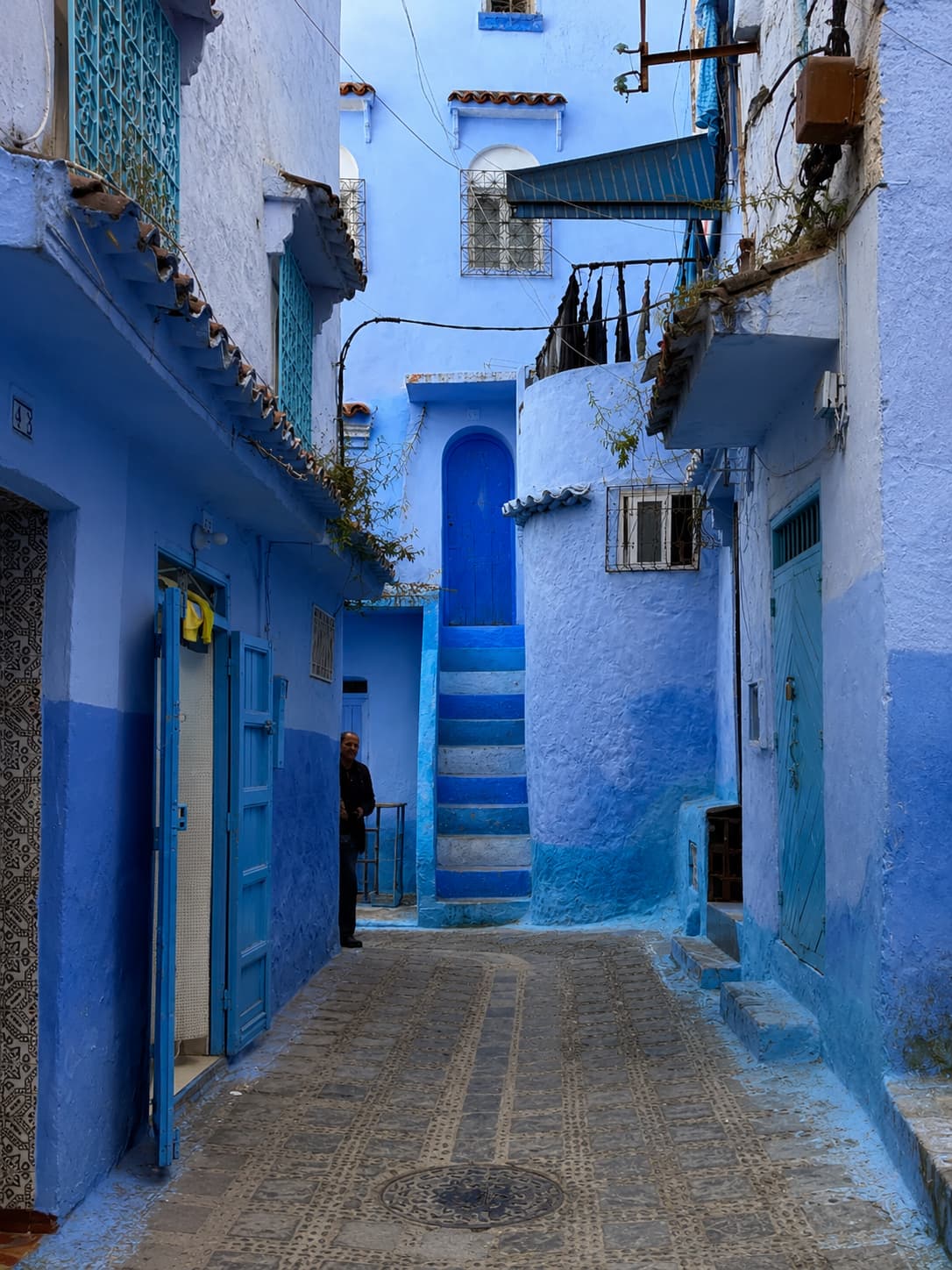 Blue-washed residential alley in Chefchaouen, Morocco, with painted stairs and a local resident standing in a doorway