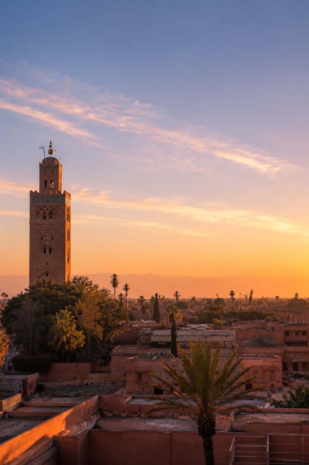 Rooftop view of the Koutoubia Mosque minaret and Marrakech skyline at sunset, warm orange light across terracotta rooftops