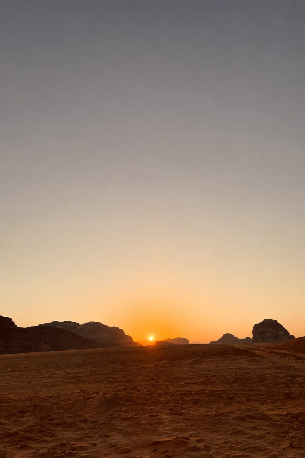 Wadi Rum desert sunset with the sun setting low between sandstone rock formations, the sky fading from orange to pale grey