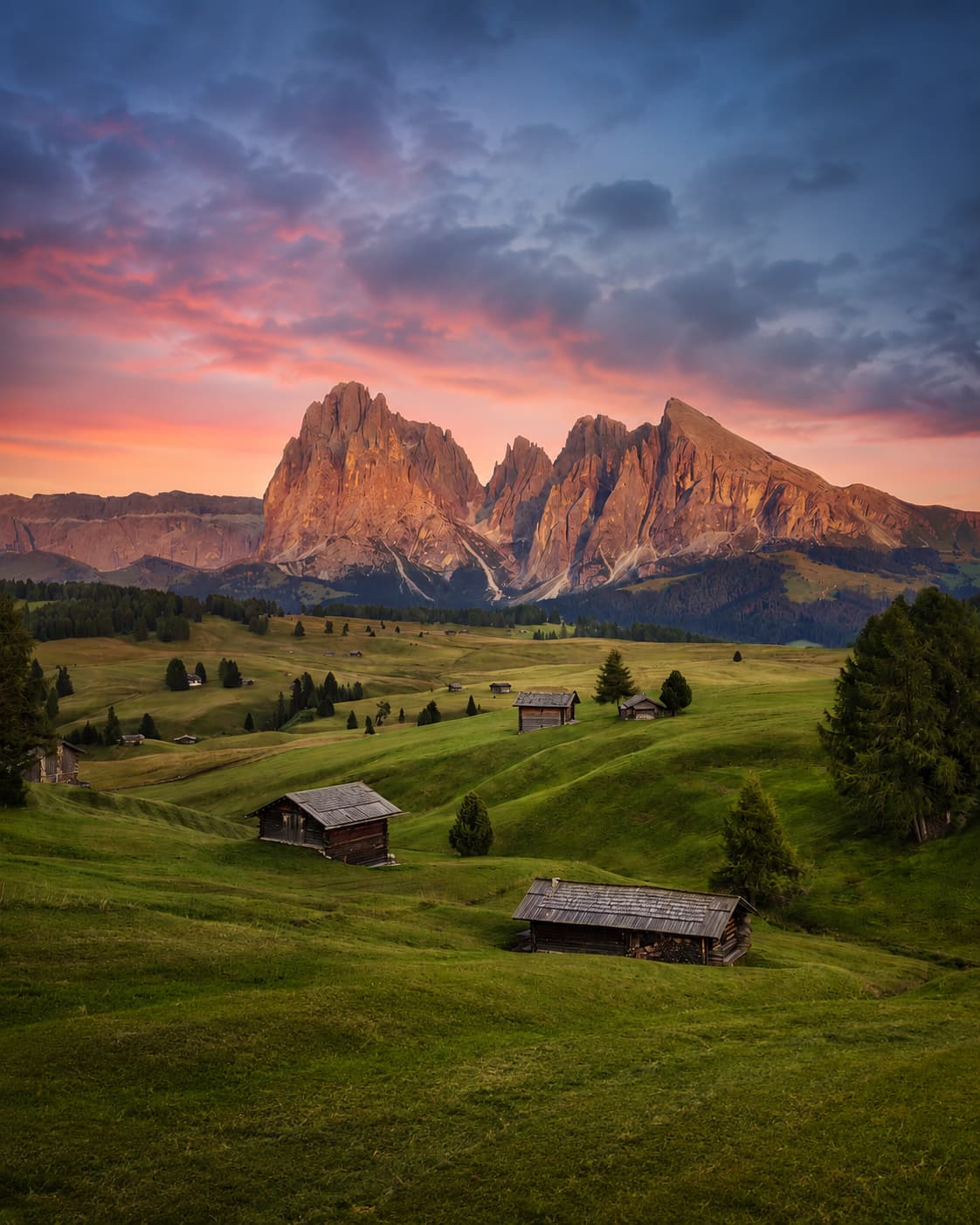 Alpe di Siusi alpine meadow at sunset with wooden mountain huts in the foreground and the Sassolungo group glowing orange-pink in a dramatic sky