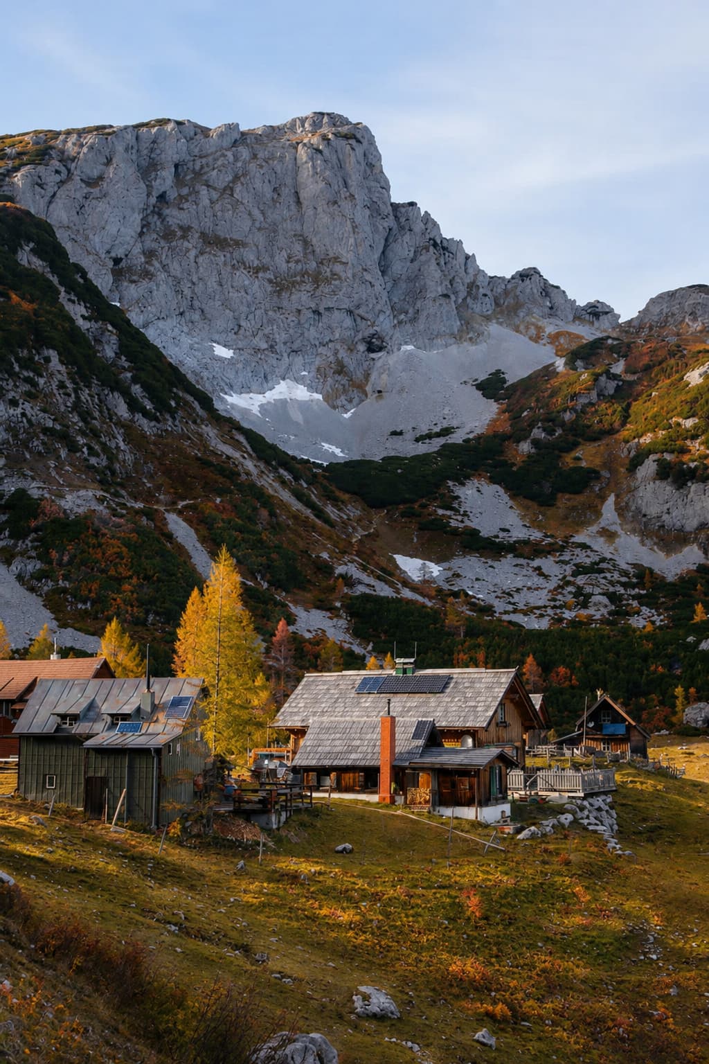 Austrian mountain huts in the alps with golden larch trees, autumn foliage, and a grey limestone cliff face dusted with early snow above