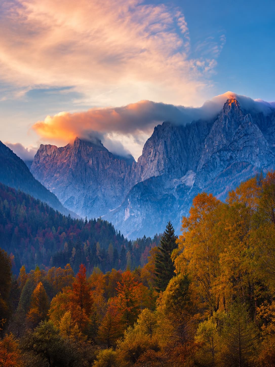 Julian Alps in Slovenia at sunset — a dramatic orange and gold autumn forest canopy below rocky grey peaks wrapped in cloud