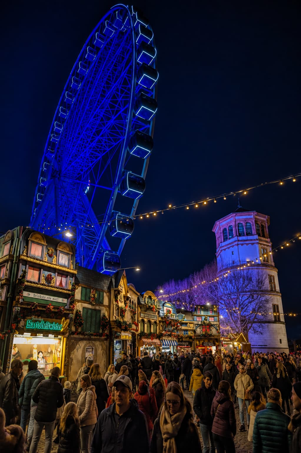 best christmas markets europe — Cologne at night with blue-lit Ferris wheel, festive stalls and medieval tower
