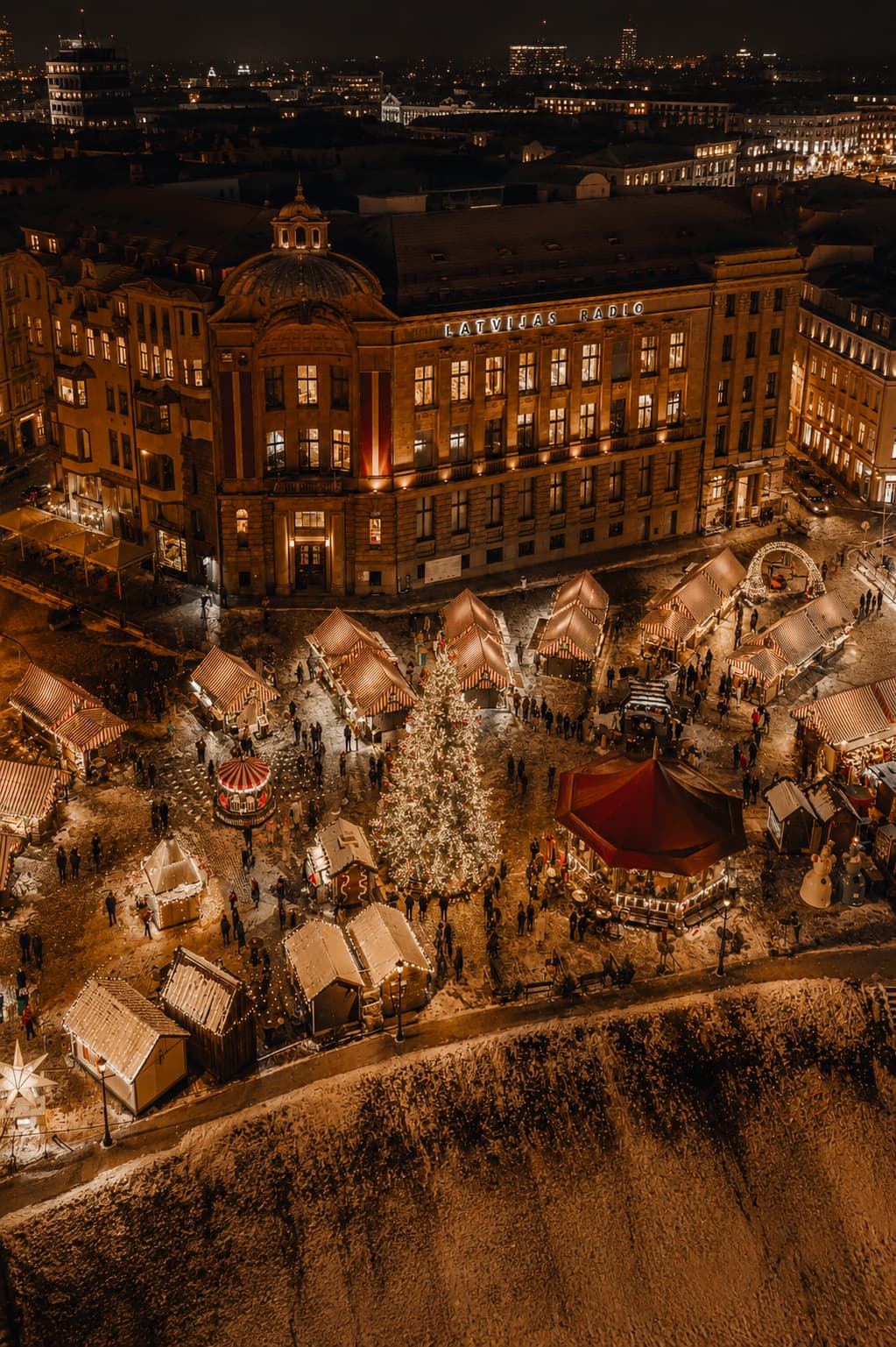 Riga Latvia Christmas market aerial at night with Latvijas Radio building, snow-covered wooden stalls around lit Christmas tree