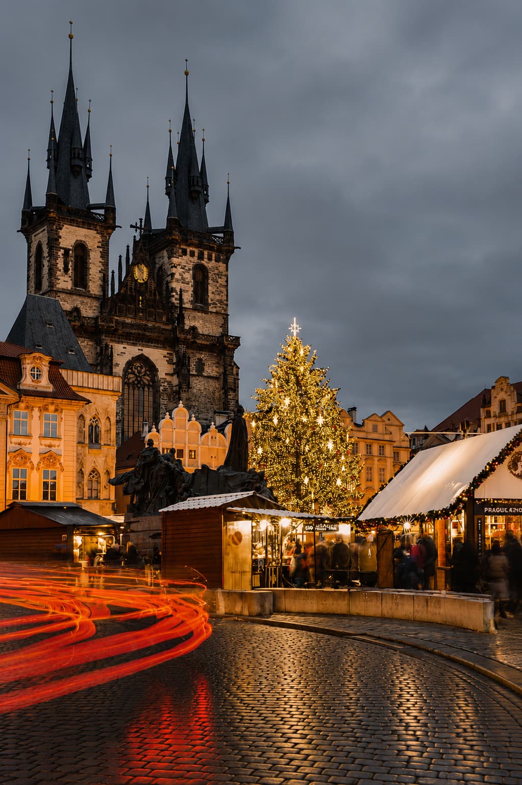 Prague Old Town Square Christmas market at night with Tyn Church spires and lit Christmas tree, long-exposure light trails on cobblestones