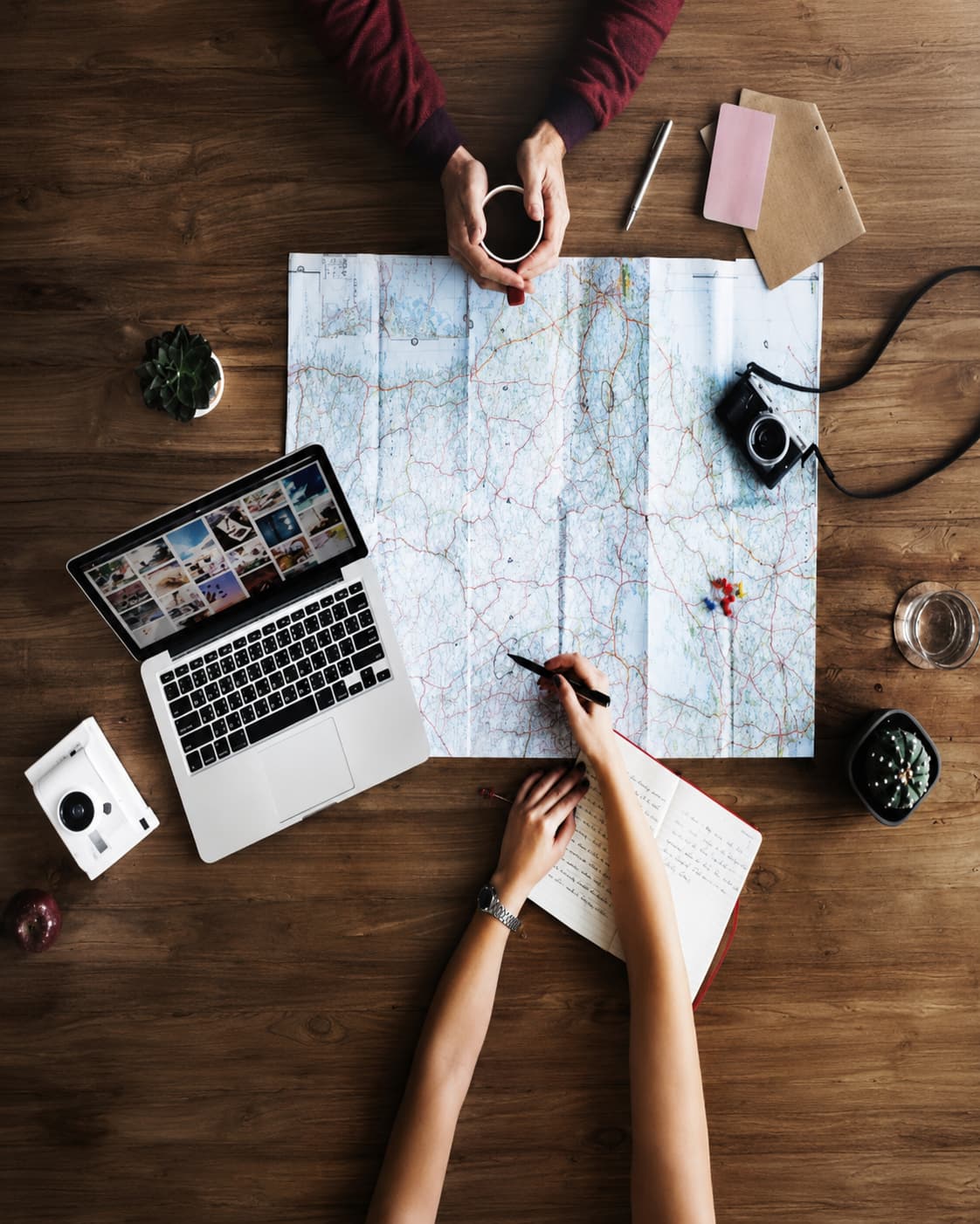 Overhead flat-lay of two people planning a birthday trip with a paper map, notebook, laptop and coffee on a wooden table