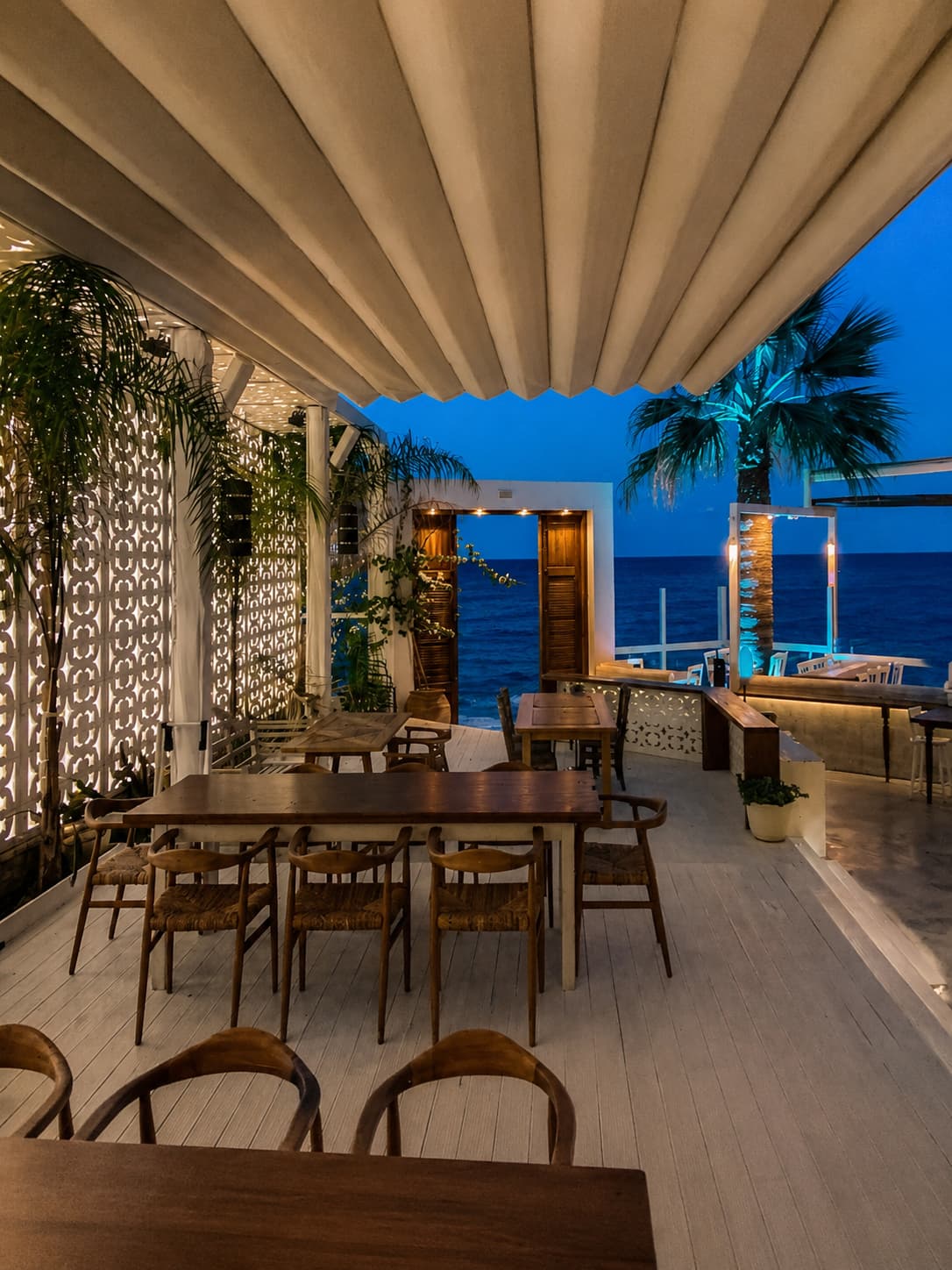 Seafront restaurant terrace in Crete at blue hour, empty dining tables, calm sea visible beyond, palm trees lit at dusk