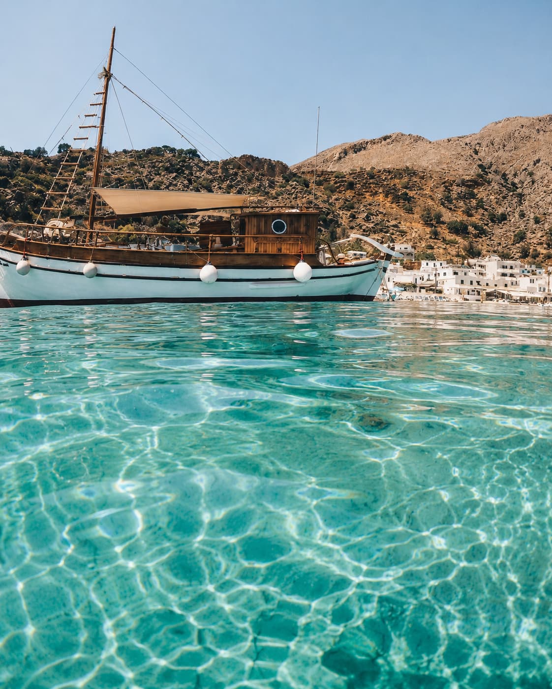 Crystal-clear turquoise water at Loutro bay on the south coast of Crete, traditional wooden sailing boat moored, white village in background