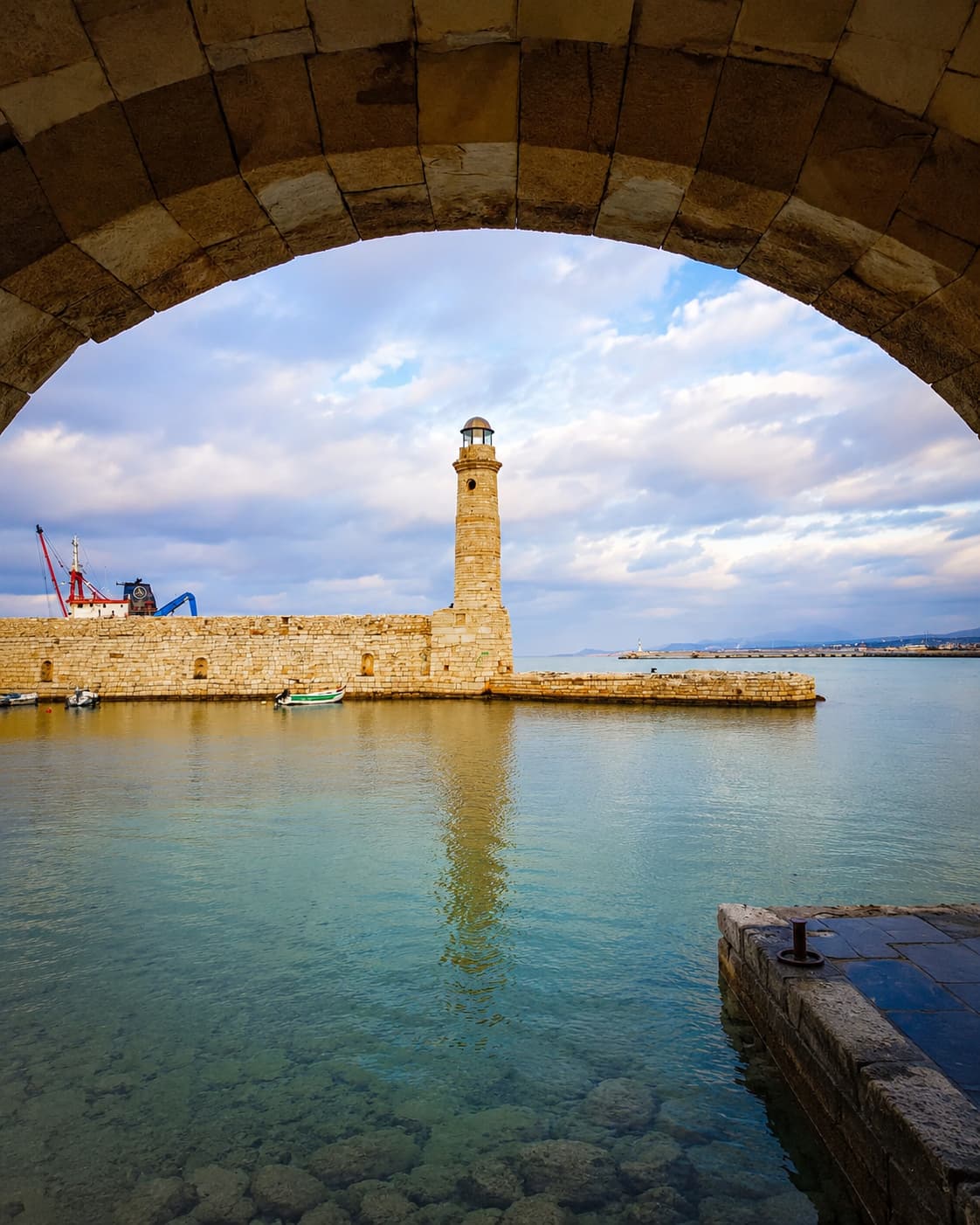 Rethymno Venetian lighthouse framed through a stone arch on Crete's north coast, turquoise water below, clouds in the October sky