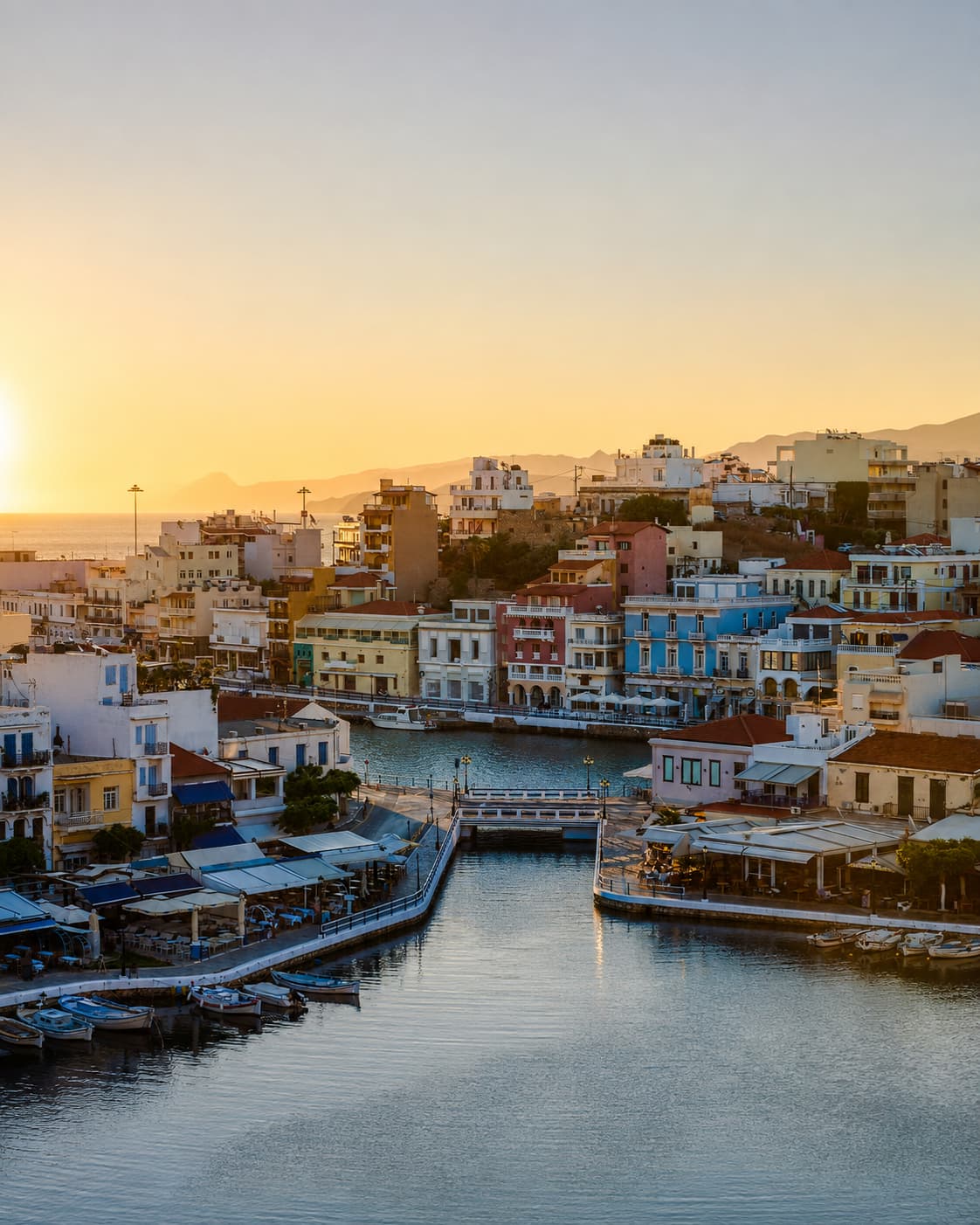 Agios Nikolaos harbour at sunset in Crete, colourful buildings reflected in the lake, golden sky, fishing boats moored along the waterfront