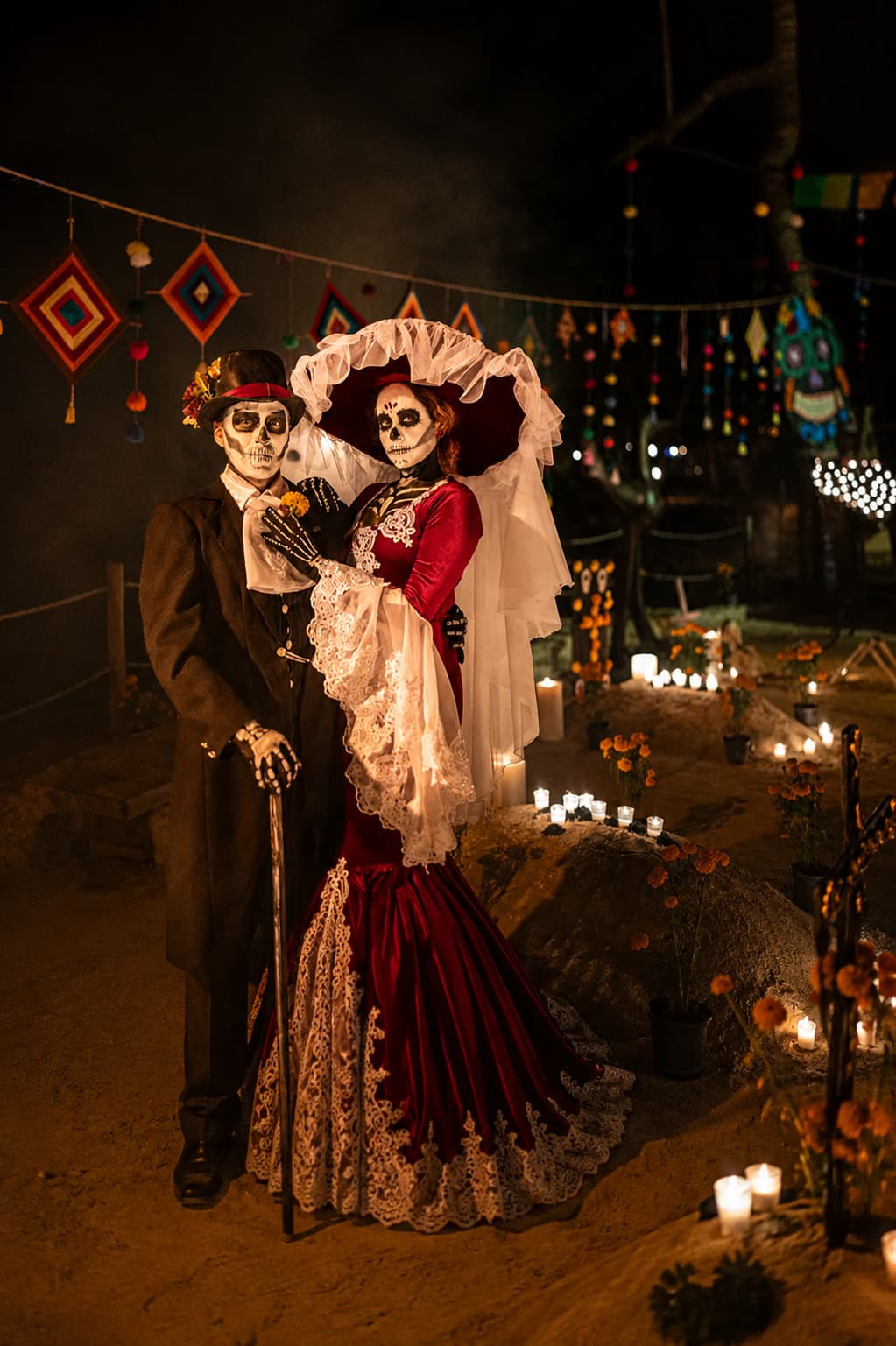 Couple in elaborate Catrina and skeleton costume at night, candles and Día de Muertos ofrenda behind them
