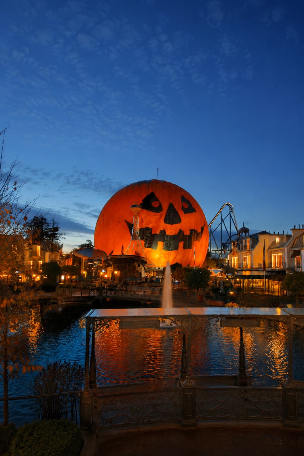 Giant illuminated jack-o-lantern balloon at a European theme park at blue hour, reflected in the water below