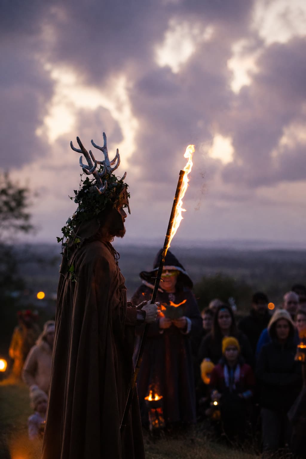 Antlered figure in ceremonial robes holding a flaming torch at a Samhain fire ceremony in Europe, crowd gathered behind at dusk