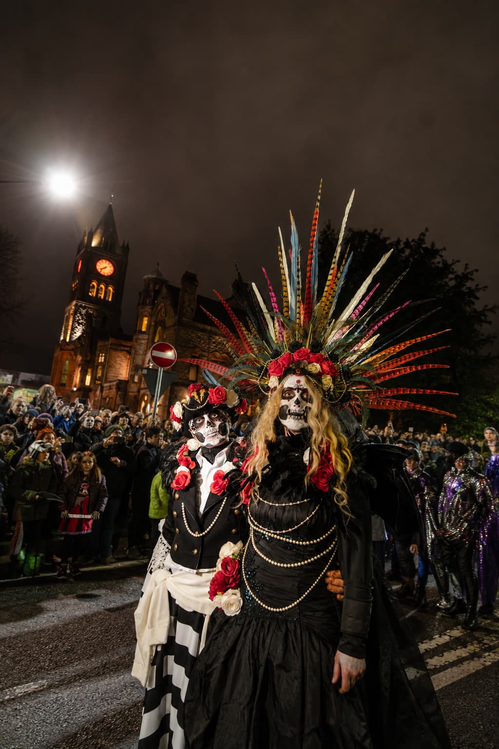 Two elaborately costumed skull-faced performers at the Derry Halloween carnival parade, historic church tower lit up behind them