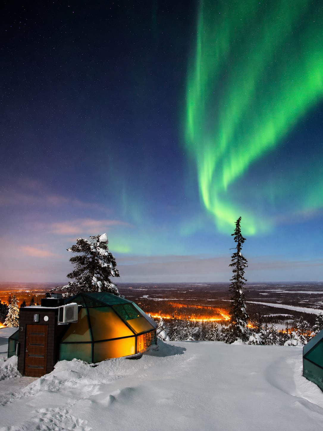 Glass igloo cabin in Lapland with bright green northern lights filling the sky above Rovaniemi — a popular accommodation choice for northern lights viewing