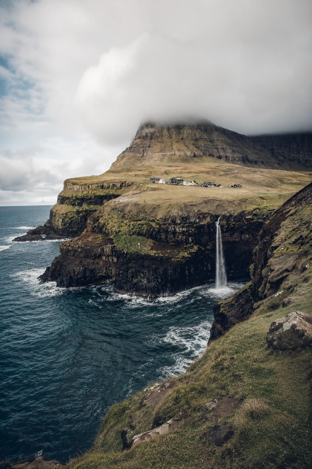 Múlafossur waterfall at Gásadalur village in the Faroe Islands, a moody travel destination with low cloud covering the clifftop mountain and ocean waves below