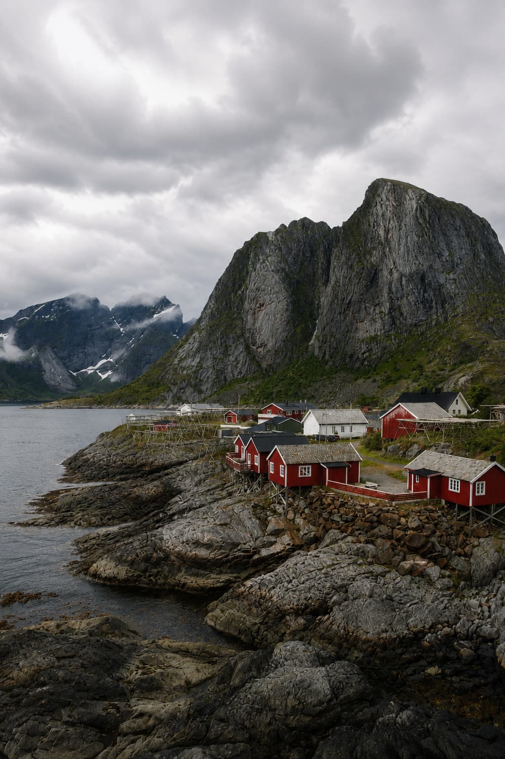 Red rorbu fishing huts at Hamnøy village in Lofoten, Norway on an atmospheric overcast day, with dramatic rocky peaks rising through cloud and snow-capped mountains behind