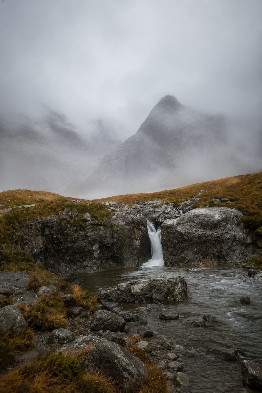 Fairy Pools on the Isle of Skye on a moody overcast day — a small waterfall cascades into a clear pool with the Cuillin Ridge summit disappearing into thick cloud behind