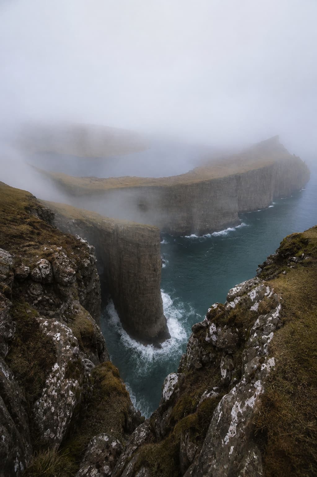 Trælanípan cliffs in the Faroe Islands in thick fog — steep sea cliffs and a sea stack partially obscured by low cloud, with turquoise Atlantic water churning far below