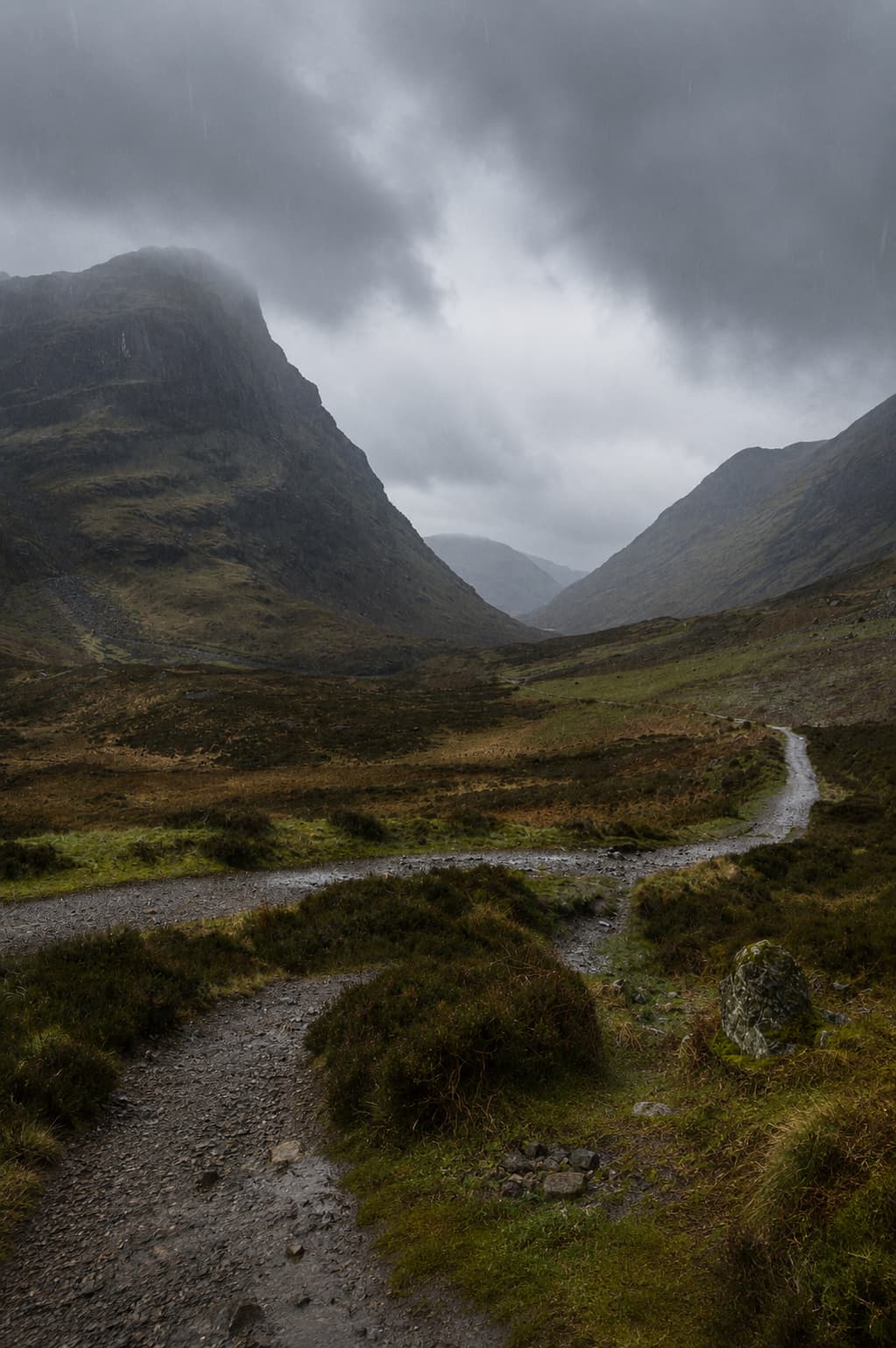 Glen Coe in the Scottish Highlands on a moody rainy day — a gravel path winds into a dramatic valley between two dark mountain walls with heavy grey cloud overhead