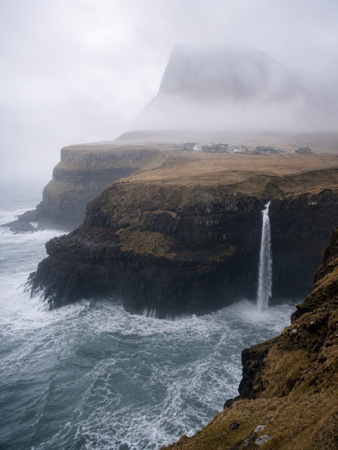 Múlafossur waterfall and Gásadalur village in the Faroe Islands on a stormy atmospheric day, with the mountain summit completely swallowed by thick fog and waves crashing below