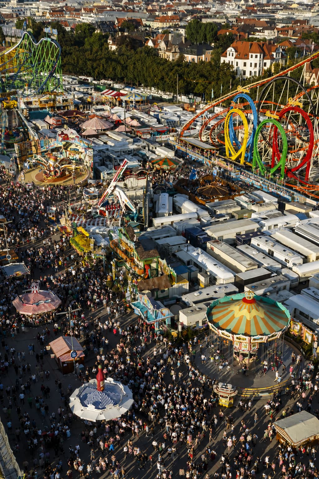 Aerial view of the Oktoberfest fairground at the Theresienwiese Munich showing rollercoasters, carousel rides, and dense crowds in golden afternoon light