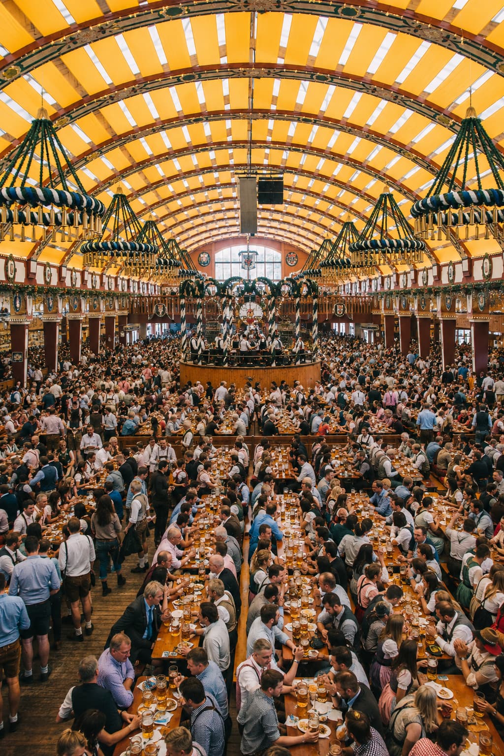 Inside a packed Oktoberfest beer tent showing long wooden benches full of visitors, golden vaulted ceiling, and a brass band on the central stage