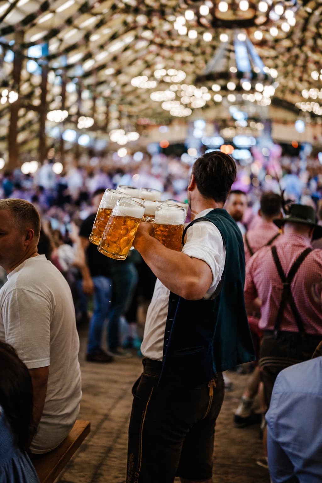 A server in traditional Lederhosen carries multiple Maß beer steins through the crowd at an Oktoberfest tent in Munich