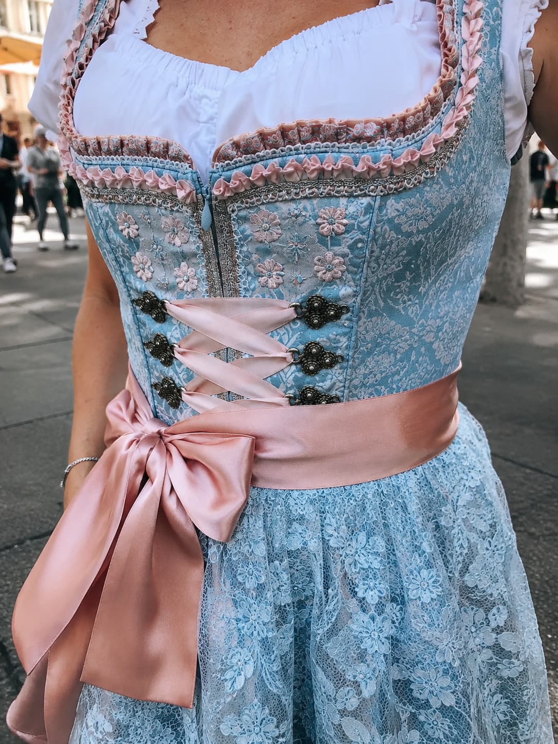 Close-up of a traditional Dirndl dress worn at Oktoberfest Munich with blue lace fabric, floral embroidery, and a pink satin ribbon bow at the waist