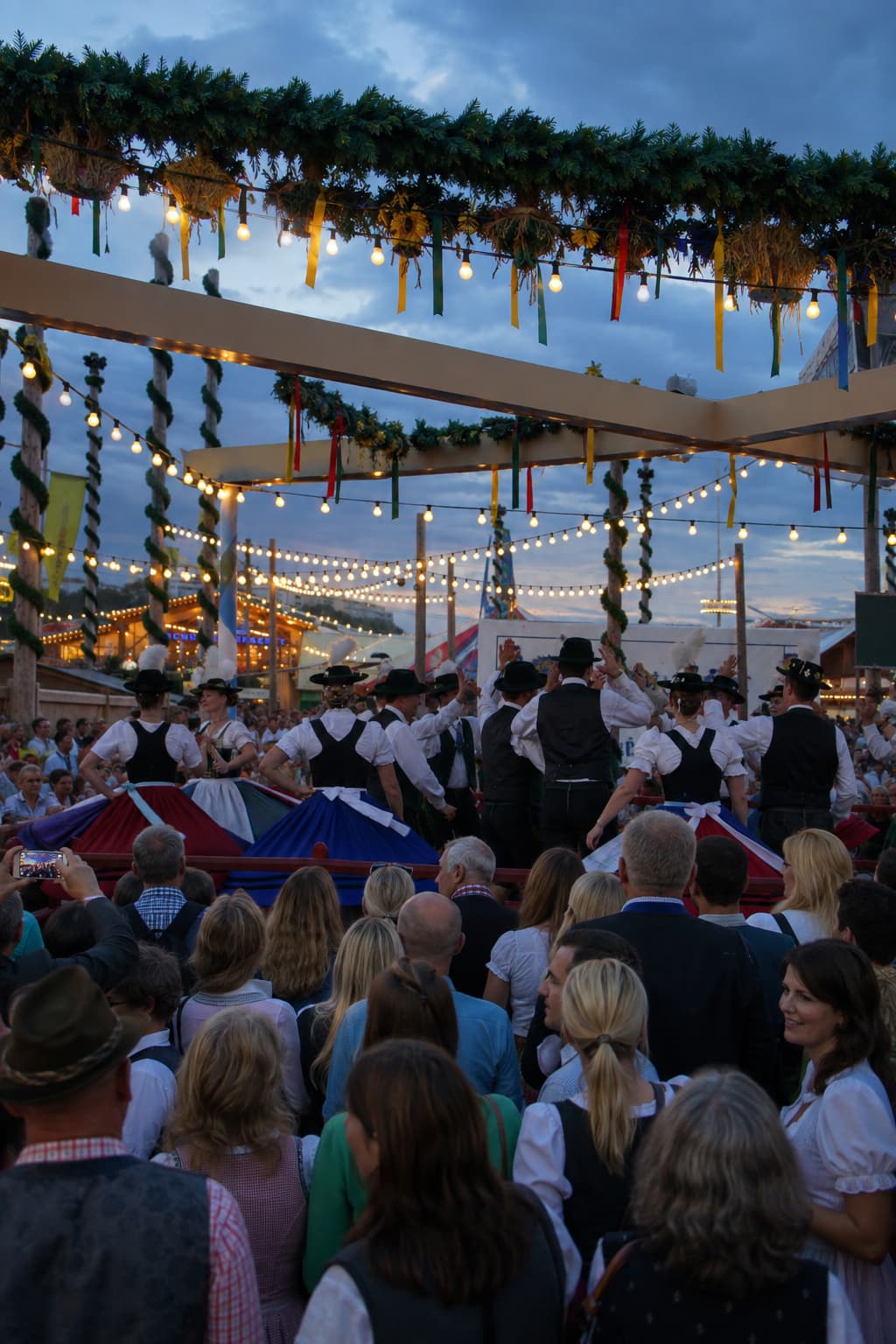 Traditional Bavarian folk dancers performing on an outdoor stage at the Oide Wiesn section of Oktoberfest Munich at dusk, with string lights and sunflower garlands overhead
