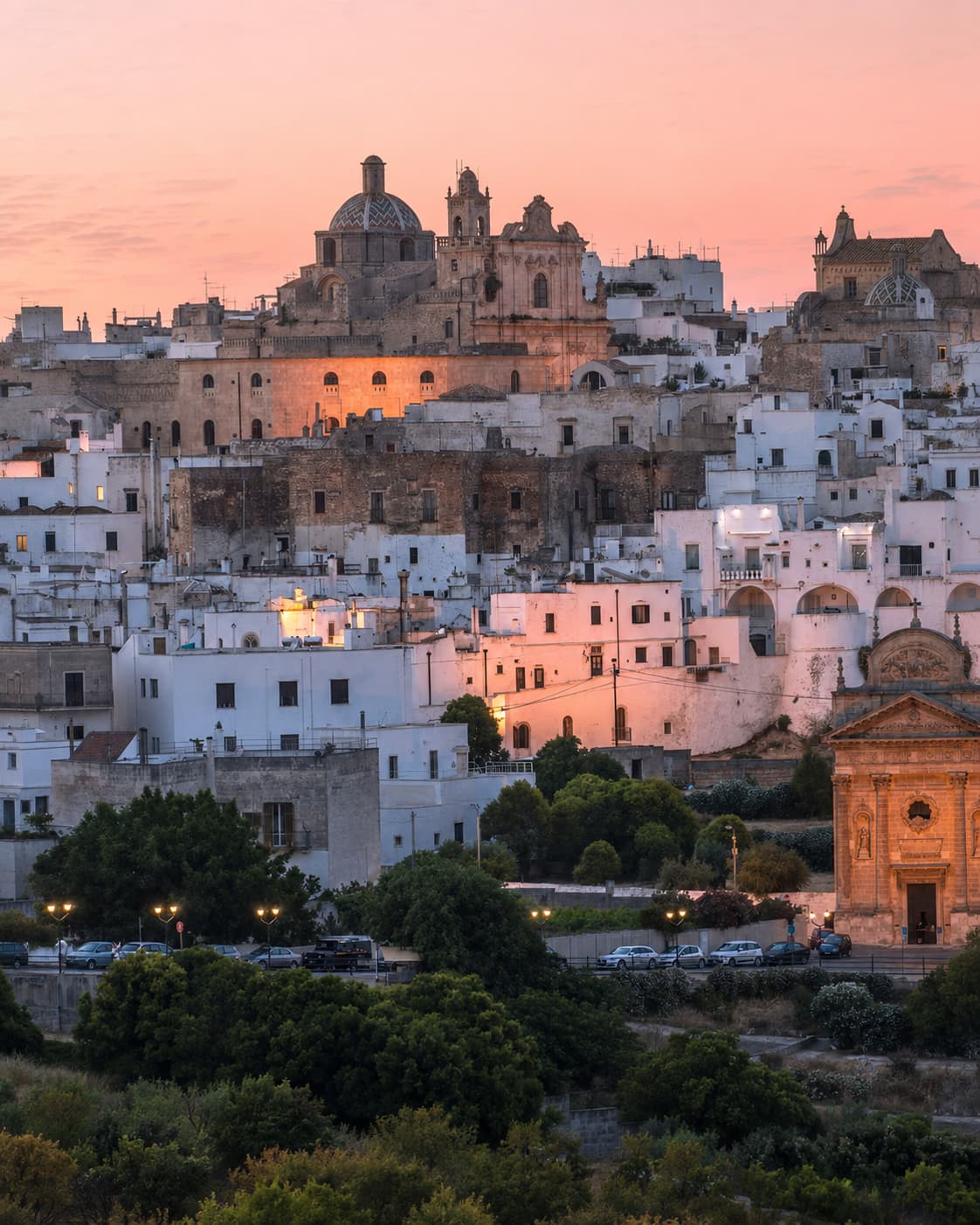 Ostuni white city at dusk in Puglia in autumn, whitewashed buildings glowing pink against a rose-coloured sunset sky, church dome and bell tower lit up