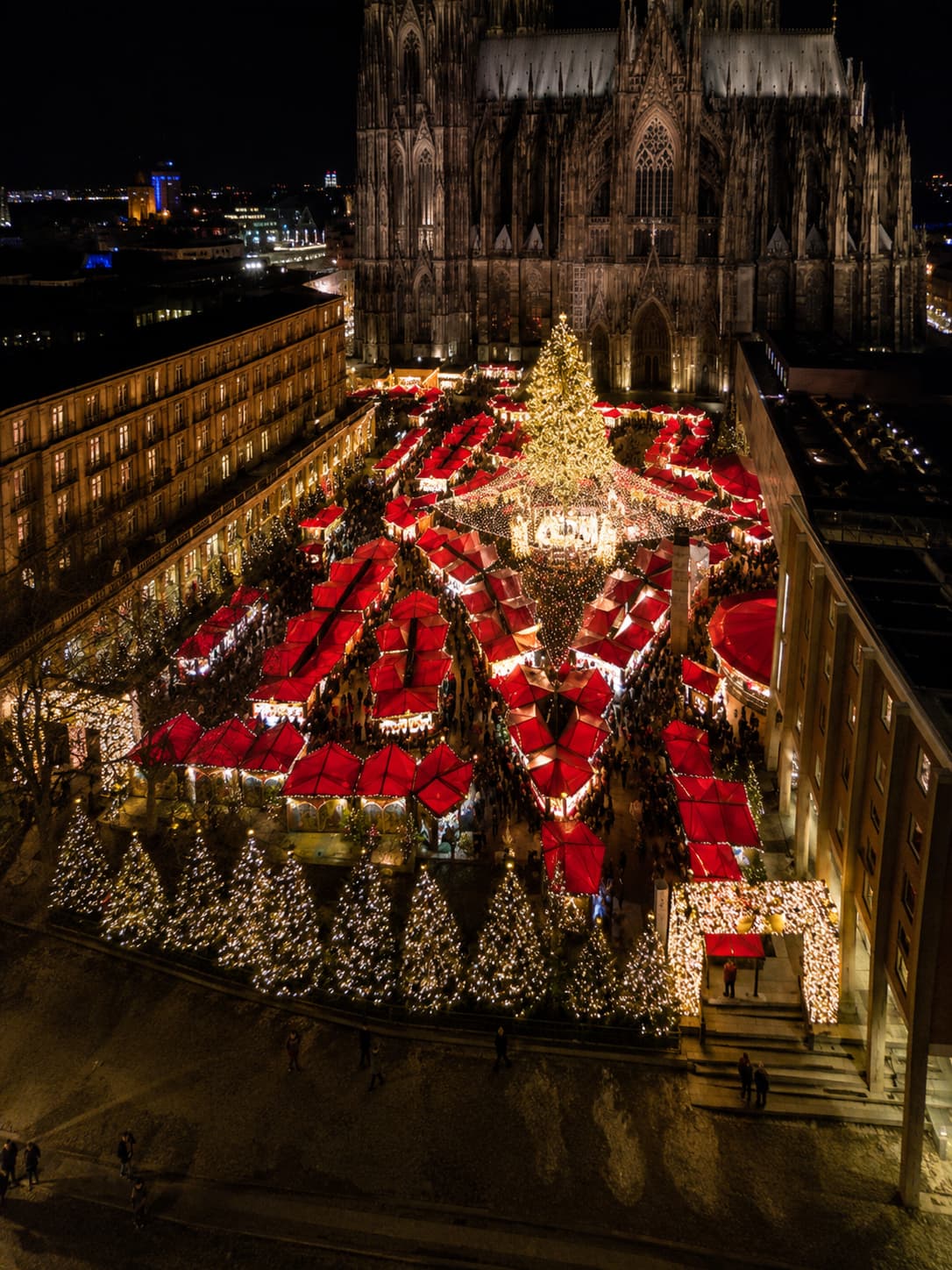 Cologne Christmas market aerial view at night with red-roofed stalls, a glowing Christmas tree and the cathedral behind — European markets open exactly during Thanksgiving travel abroad week in 2026