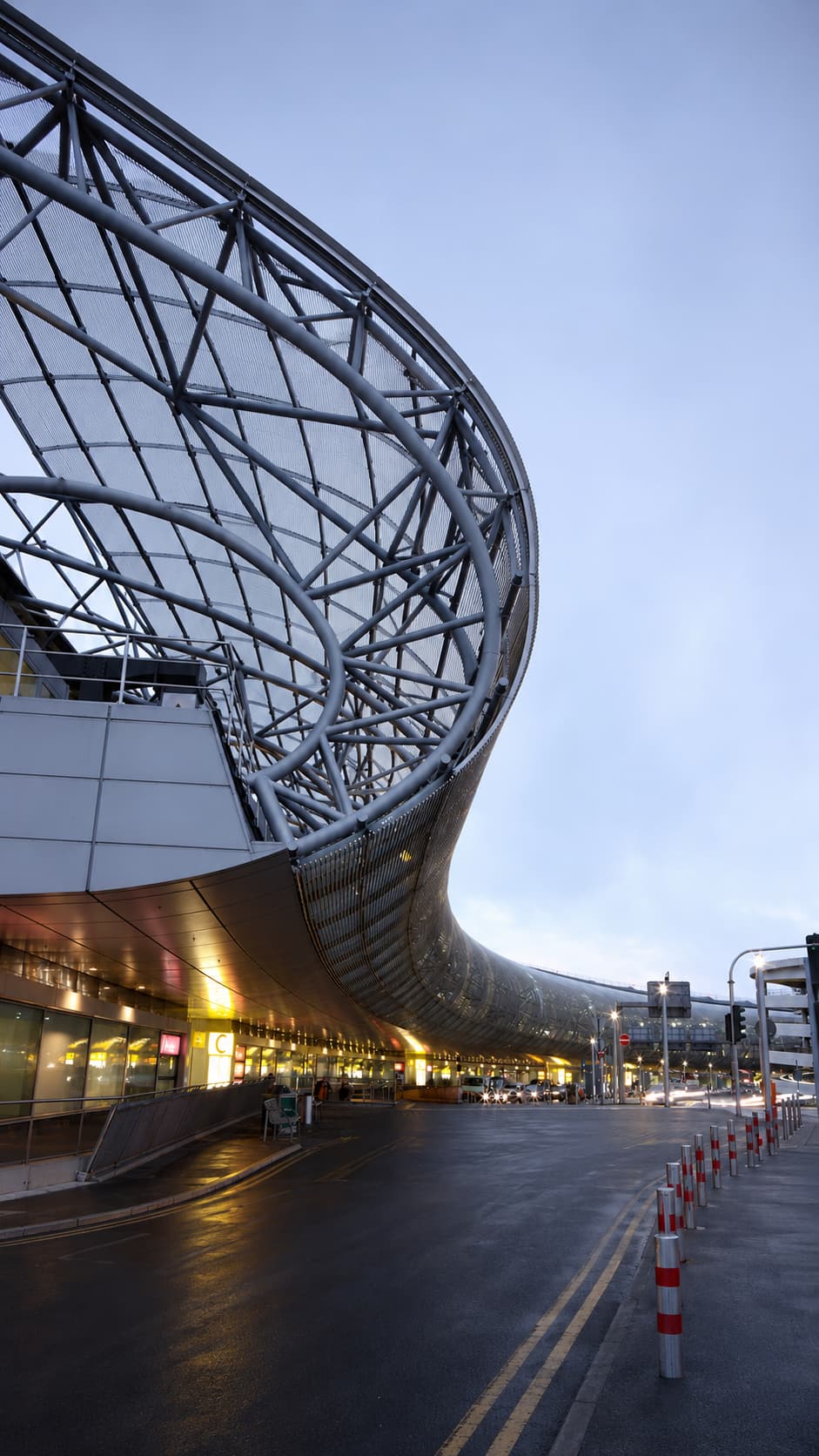 Modern airport terminal exterior at dusk with curved steel and glass architecture lit from within — the international terminal is notably quieter than domestic gates during Thanksgiving week