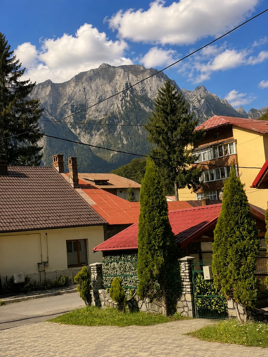 Piatra Craiului limestone peaks rising above Zărnești village rooftops in Transylvania — red-tiled houses, conifers, and a clear blue sky on the drive from Bucharest