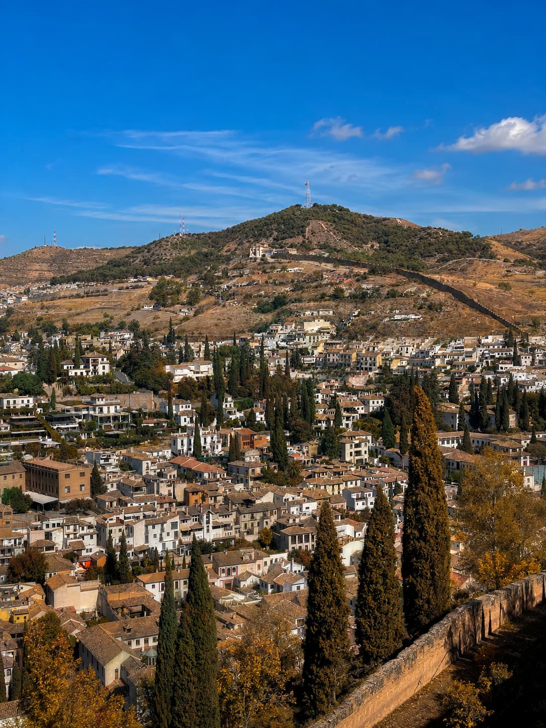 Granada Albaicín neighbourhood in Andalusia in autumn — warm October foliage and cypress trees above hilltop whitewashed houses against a blue sky