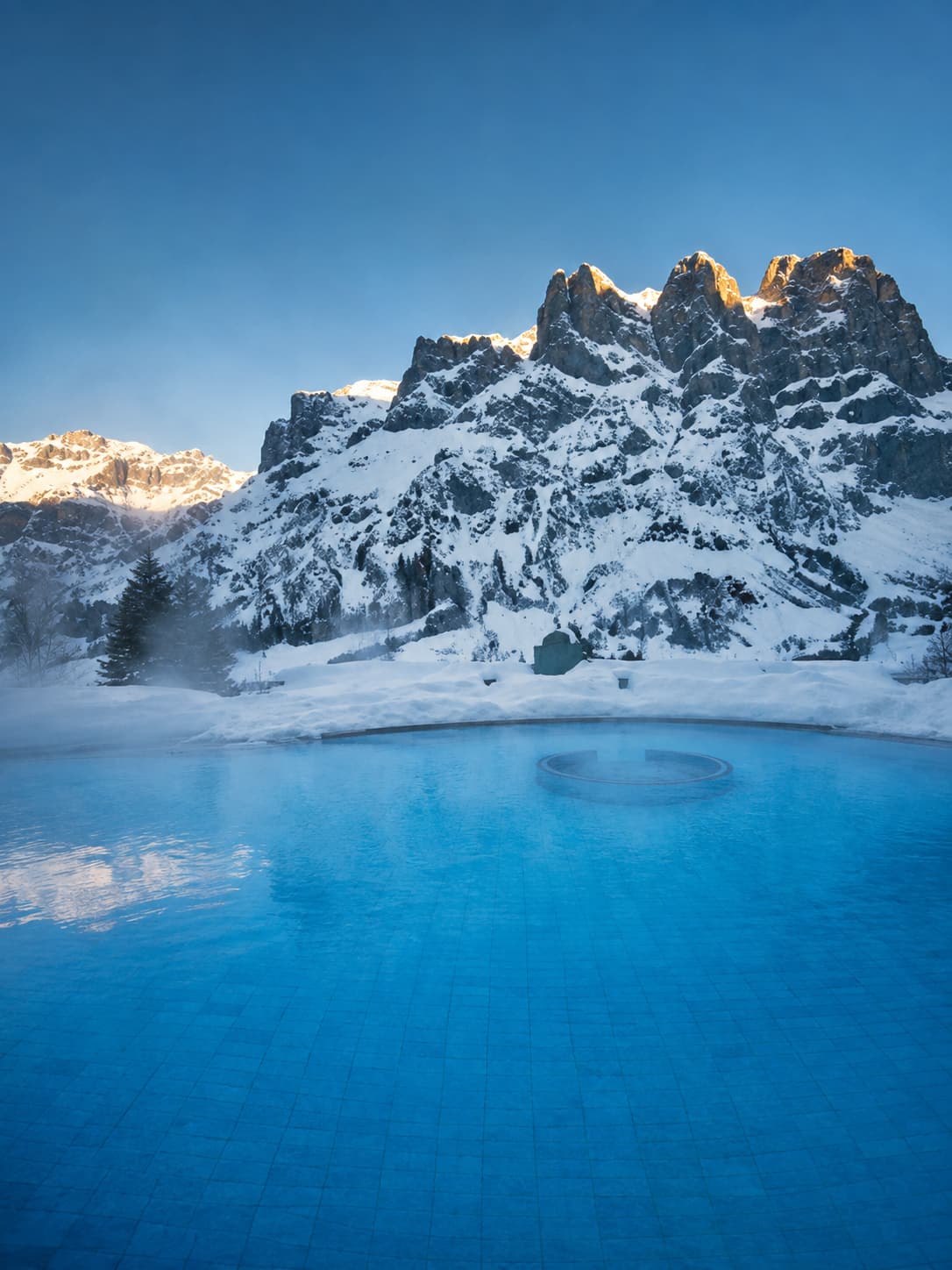 Outdoor thermal pool at Leukerbad, Switzerland — the highest thermal spa town in Europe — with steam rising from the heated water and snow-covered alpine peaks of the Gemmi Pass rising directly behind