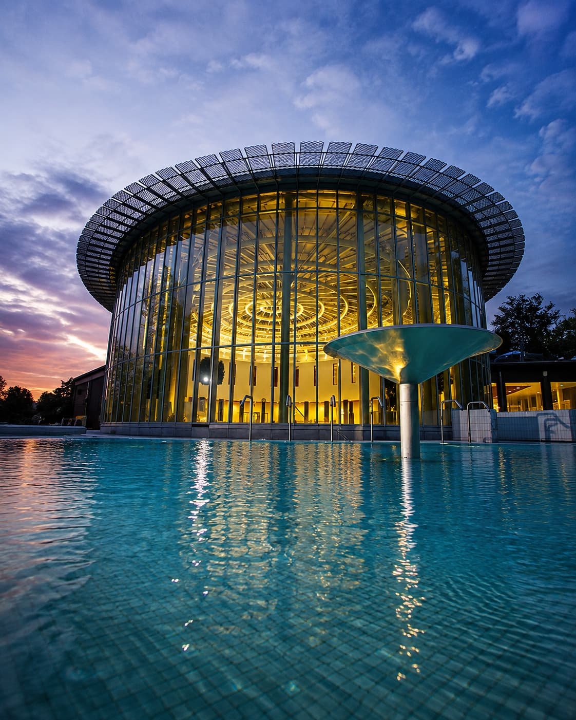 The Caracalla Terme spa building in Baden-Baden illuminated at dusk, with its circular glass facade reflected in the outdoor thermal pool — a modern counterpart to the historic European spa towns