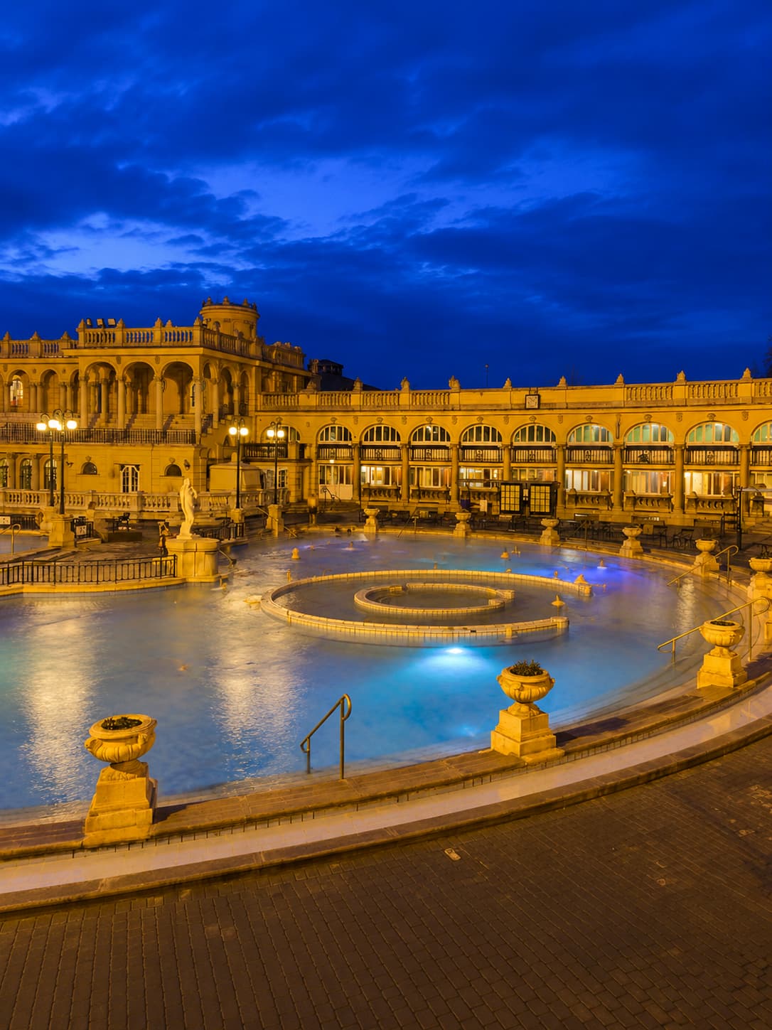 Széchenyi thermal bath in Budapest at night — the outdoor pool of one of Europe's most spectacular thermal bath cities lit in gold and blue, with the Neo-Baroque facade rising behind the steaming water